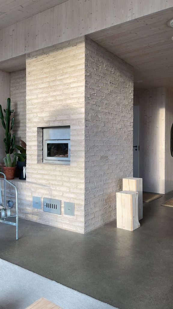 Studio Takkusen kohteessa suuri vaalea tulisija jonka syvennyksessä viherkasveja ja seinustalla puisia pölkkyjä